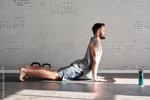 Muscular athlete tattooed body exercising workout in sunny sport club, brick wall. Fit shirtless male fitness model.