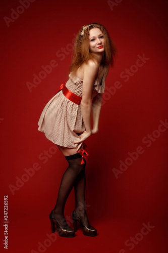 Shy prissy pretty young woman in short dress, red belt, black stockings with red ribbons and high heels fixes her dress against the red background. Pin-up full-growth retro-style portrait