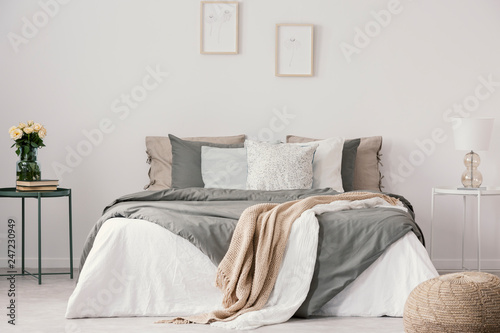 Flowers on table next to bed with pillows in white bedroom interior with posters and pouf. Real photo