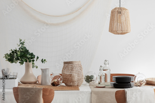 Green plant in beige vase, white jug and wicker lantern on dining table in bright interior photo