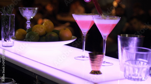 The bartender in two beautiful glasses, half full, squeezes the next ingredient, adds whipped foam, glasses at the bar are near the pizza with citrus fruits
