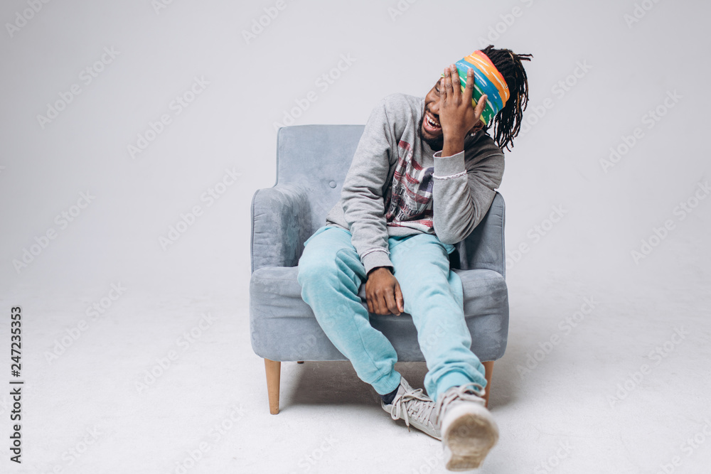 Portrait of a smiling black young model in a gray sweater and blue pants on a gray background near the chair Portrait of a smiling black young model in a gray sweater and blue pants on a gray background near the chair