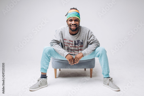 Portrait of a smiling black young model in a gray sweater and blue pants on a gray background near the chair