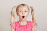 Closeup portrait of surprised white blonde Caucasian preschool girl making faces in front of camera. Child smiling laughing posing in studio on plain light background. Kid expressing emotions