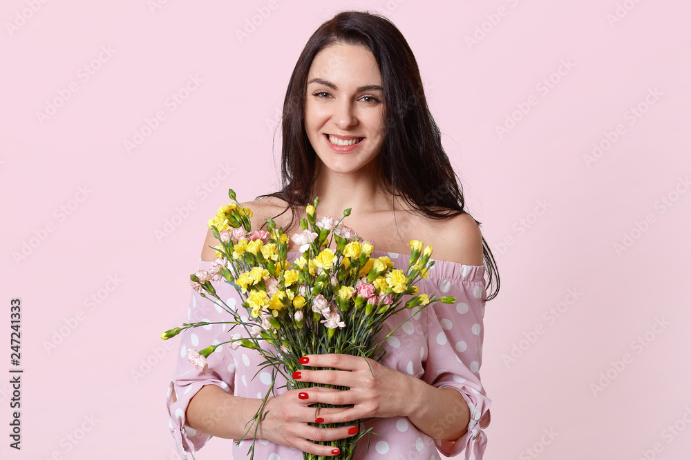 Happy positive woman with dark hair, holds flowers in hands, smiles positively, enjoys spring warm day, dressed in stylish polka dot dress, isolated over pink background. Ladies like flowers