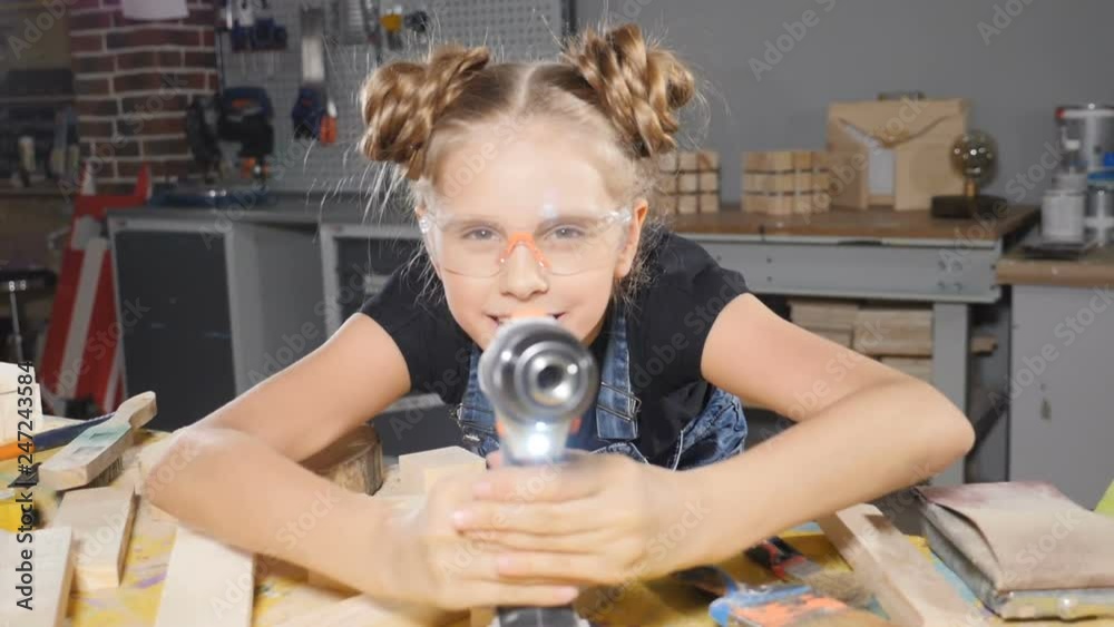 Portrait of funny 10 year old girl in wood carpentry holding an electronic drill, posing at camera. Little builder concept. hd Portrait of funny 10 year old girl in wood carpentry holding an electronic drill, posing at camera. Little builder concept. hd