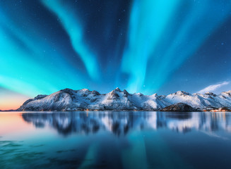 Northern lights and snow covered mountains in Lofoten islands, Norway. Aurora borealis. Starry sky with polar lights and snowy rocks reflected in water. Night winter landscape with aurora, sea. Nature