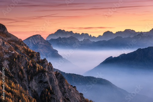 Mountains in fog at beautiful sunset in autumn in Dolomites  Italy. Landscape with alpine mountain valley  low clouds  trees on hills  orange sky with clouds at dusk. Aerial view. Passo Giau. Nature