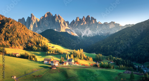 Fototapeta Naklejka Na Ścianę i Meble -  Landscape with village with houses, church, green meadows, trees, rocks, blue sky. Santa Maddalena in mountain valley at sunset in autumn in Dolomites, Italy. St. Magdalena and mountains. Val di Funes