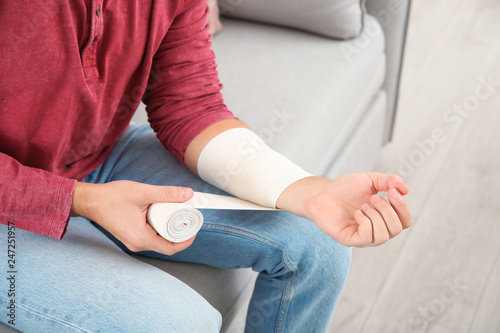 Young man applying bandage on injured arm at home, closeup. First aid