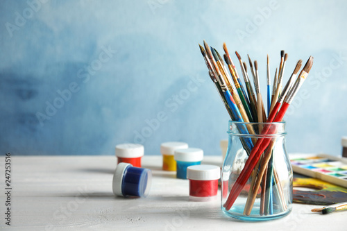 Glass jar with brushes and paints on table against color background. Space for text
