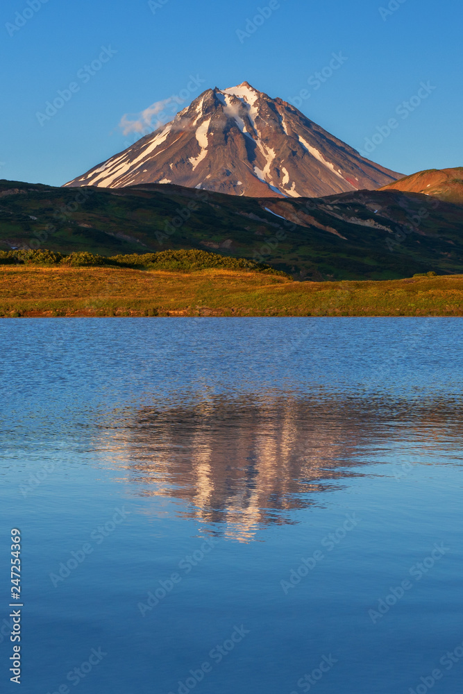 Stunning autumn volcano landscape at sunset: scenery evening view of cone volcano and reflection of mountains in picturesque mountain lake. Kamchatka Peninsula, Russian Far East, Eurasia.