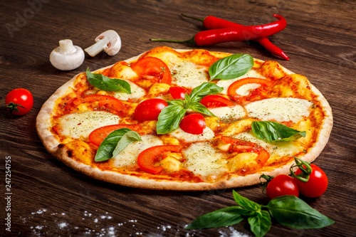 Pizza Italian margherita with melted mozzarella cheese, red cherry tomatoes and fresh green basil leaves on a brown table decorated by mushrooms, red sweet pepper and cherry tomatoes