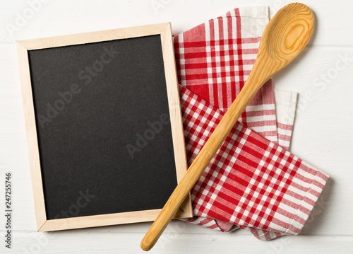 Blank, empty, black chalkboard with wooden cooking spoons and red checkered dish towel flat lay from above on white wooden table
