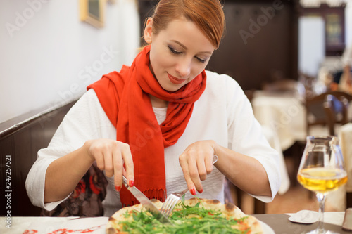 Smiling woman eating pizza with arugula in italian restaurant. Irish model in white blouse sweater clothing, red scarf, redhead hair sitting at table in pizzeria background.