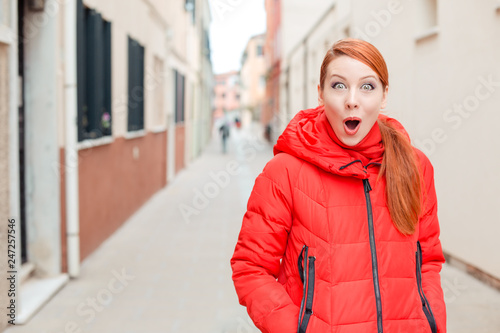Deal or sale, shocking news. Shocked woman in Murano, Venice, Italy. Irish model in red winter coat clothing, redhead hair standing on urban background. Amazed, stunned, emotion, expression concept. 