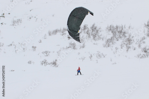 Professional kite boarding rider sportsman with kite in sky rides in snow  on kiteboard. Recreational activity, extreme active sports, snowkiting ski snowboard, outdoor, winter snowy season