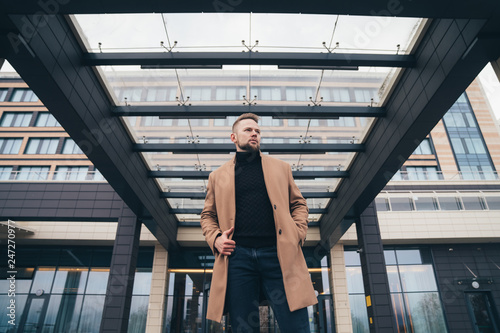 Attractive man in trendy brown coat posing near business center. Handsome european man with beard looks away. Concept lifestyle, fashionable men style