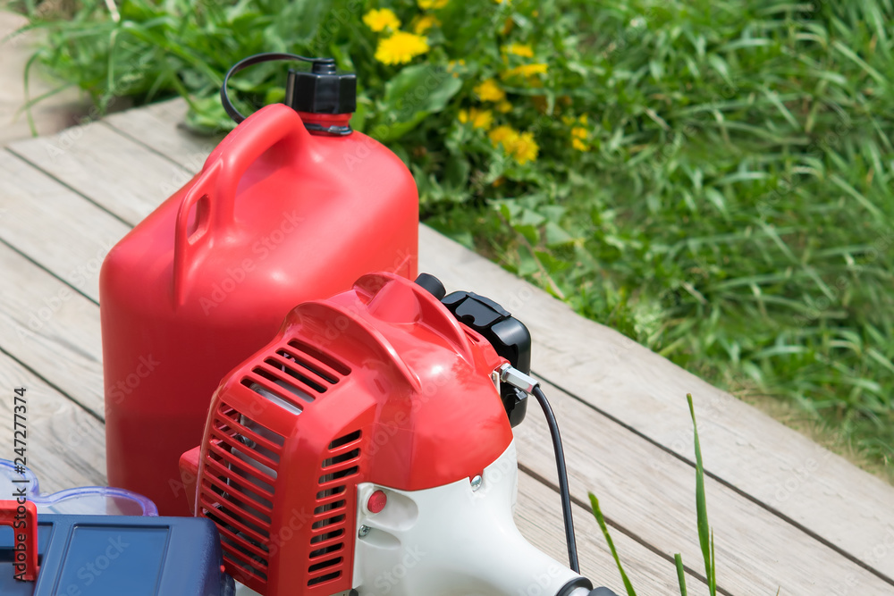 Red lawn mower and fuel canister lie on a wooden table against a green lawn Red lawn mower and fuel canister lie on a wooden table against a green lawn