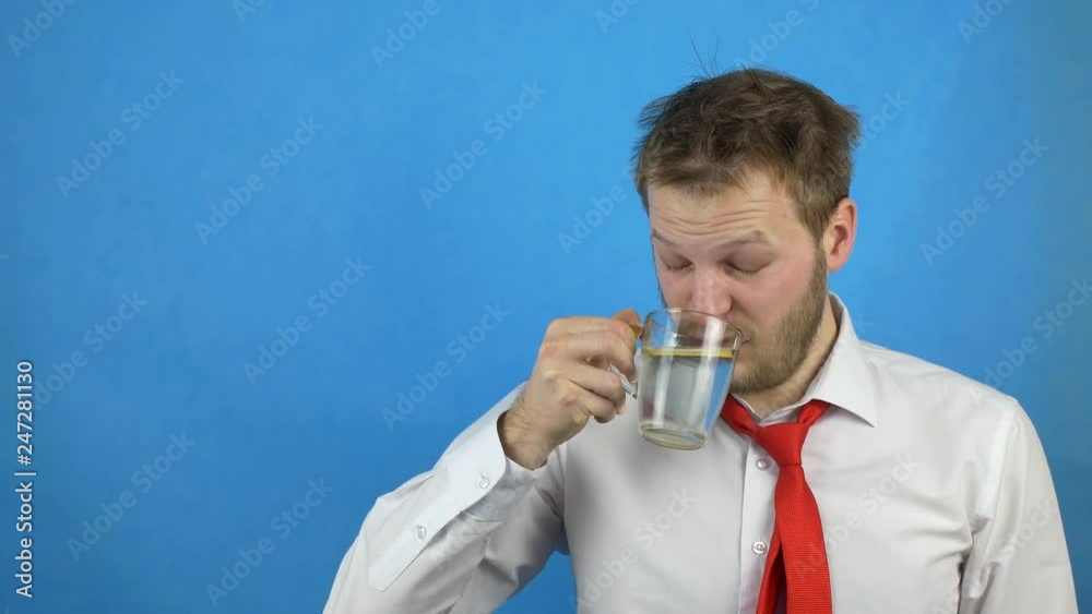 Young caucasian male businessman in white shirt and hangover tie drinks water with lemon, drowsiness, blue background, copy space Young caucasian male businessman in white shirt and hangover tie drinks water with lemon, drowsiness, blue background, copy space