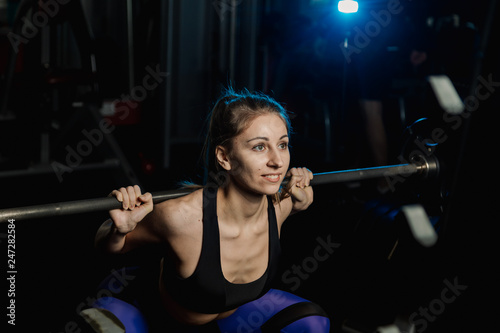 Active beautiful fitness model girl crouches with a barbell on the shoulders in the gym.