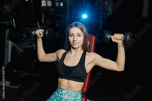 Active beautiful fitness model girl trains with dumbbells on the bench in the gym.