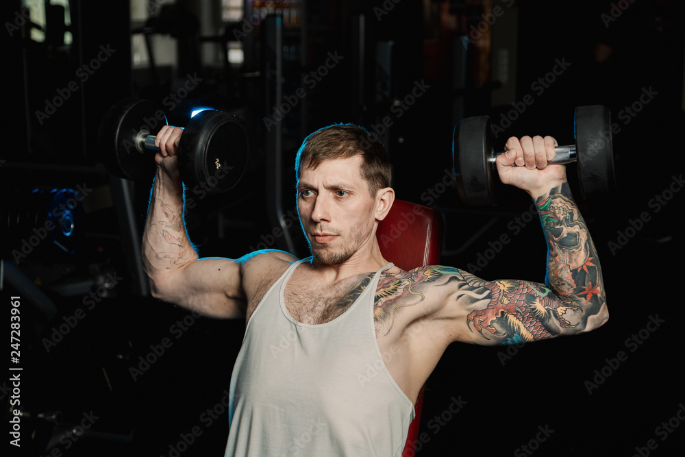 Handsome athletic man training his shoulders with dumbbells sitting on a bench in the gym. Handsome athletic man training his shoulders with dumbbells sitting on a bench in the gym.