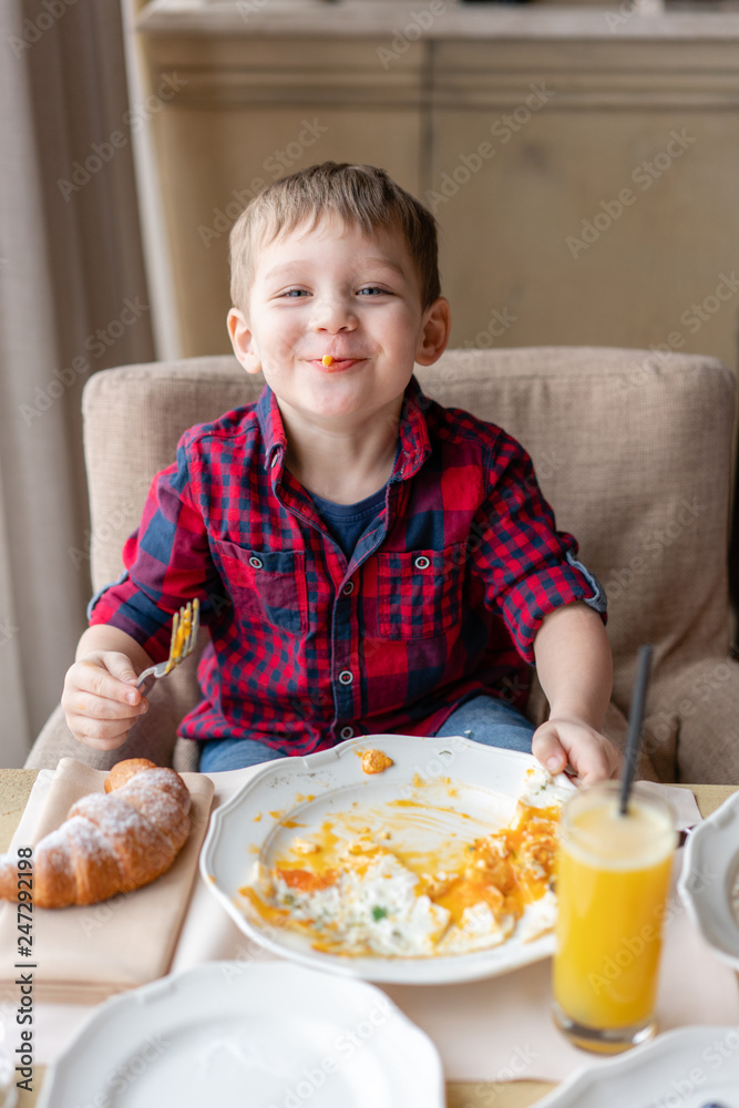 Funny and happy boy having Breakfast. Light brunch near window in a cafe. Croissants, omelet, orange juice and many different dishes on the table in the cafe. Funny and happy boy having Breakfast. Light brunch near window in a cafe. Croissants, omelet, orange juice and many different dishes on the table in the cafe.