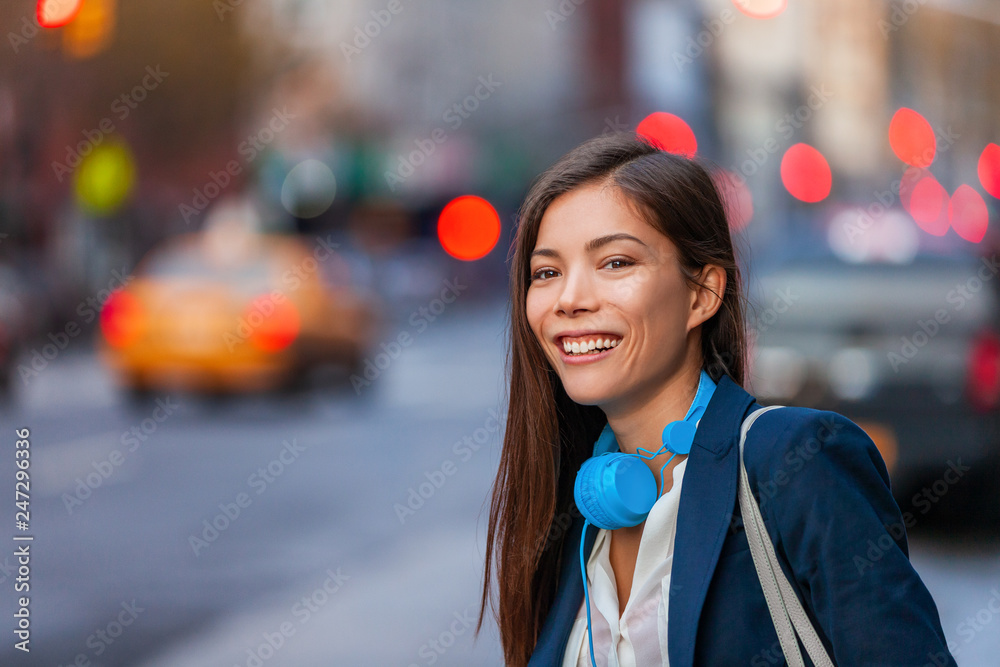 Fototapeta premium Young professional Asian woman hip with tech device headphones walking on NYC New York city street commuting after work at night. Happy smiling biracial chinese young girl urban lifestyle.
