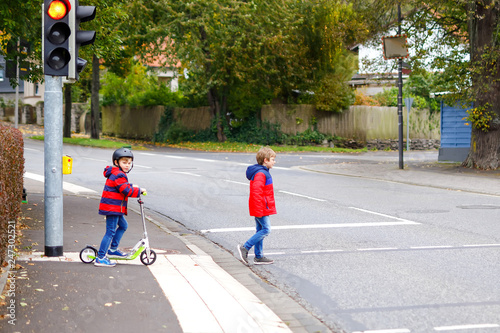 Two little schoolkids boys running and driving on scooter on autumn day. Happy children in colorful clothes and city traffic crossing pedestrian crosswalk with traffic lights. photo