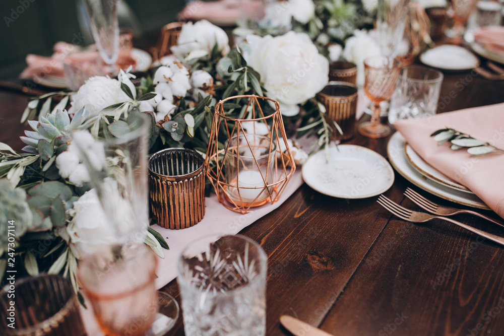on wooden banquet table are glasses, plates, candles, table is decorated with compositions of cotton and eucalyptus branches, plates are decorated with napkins and sprig of Italian greenery - obrazy, fototapety, plakaty 