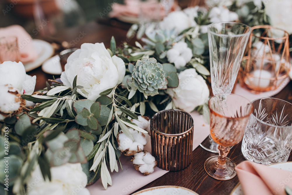 on wooden banquet table are glasses, plates, candles, table is decorated with compositions of cotton and eucalyptus branches - obrazy, fototapety, plakaty 