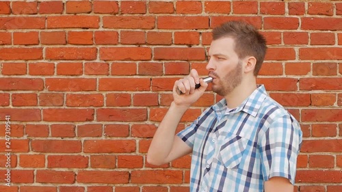 Fashionable and stylish guy with a beard smoking a vape against a brick wall, copy space, slow-mo, outside photo