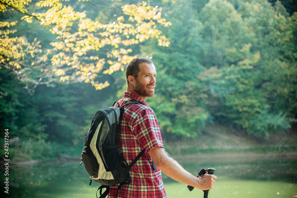 Happy young man in red checkered shirt standing by the mountain lake surrounded by lush green forest. Looking over his shoulder and smiling