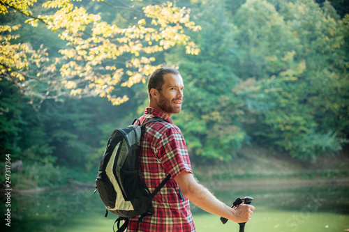 Obraz na płótnie Happy young man in red checkered shirt standing by the mountain lake surrounded by lush green forest. Looking over his shoulder and smiling w salonie