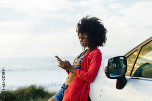 Fashionable afro hair woman on vacation texting on smartphone towards the sea. Stylish black model relaxing on a car trip to the coast.