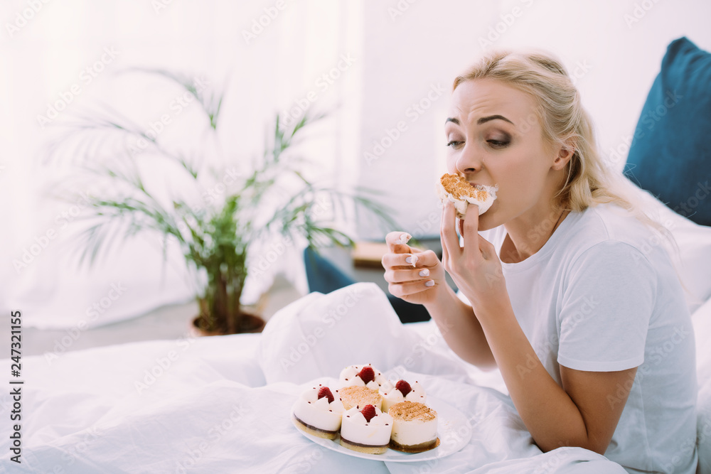 selective focus of stressed woman in pajamas eating cake in bed alone