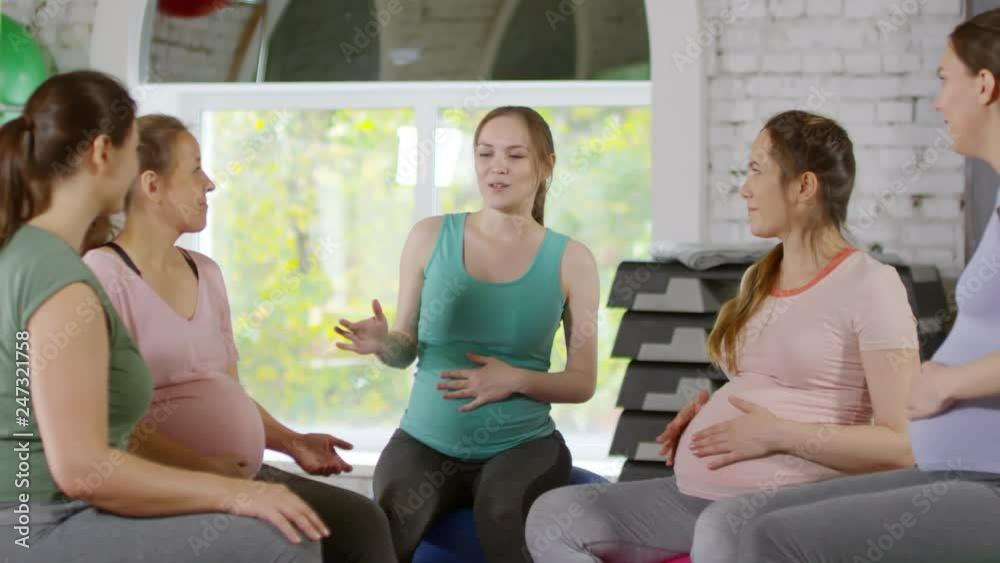 Group of five cheerful pregnant women sharing experience and talking while sitting on stability balls in gym