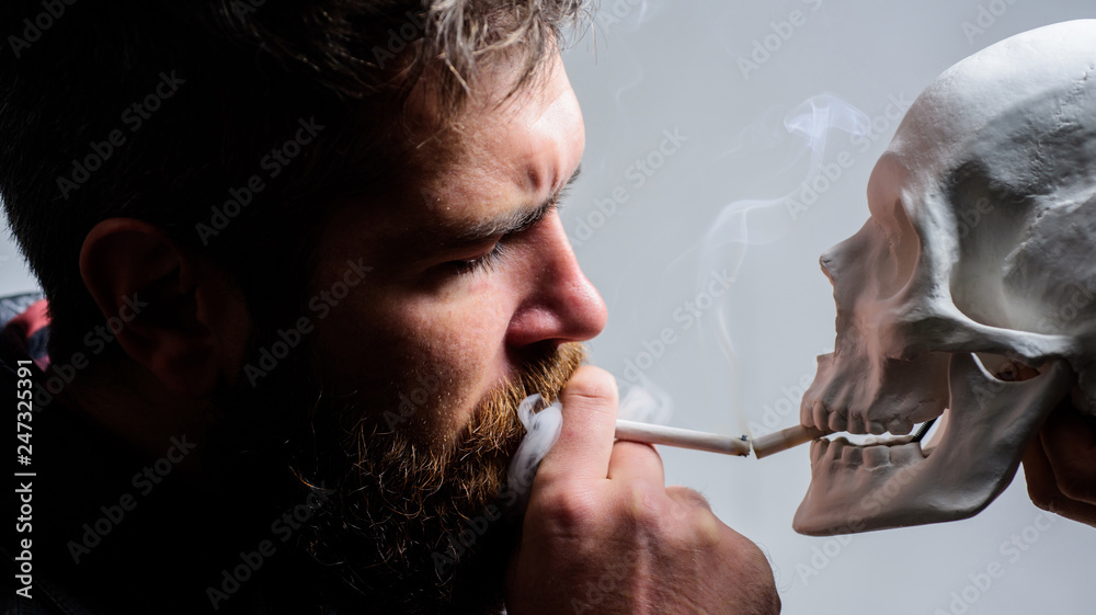 Fototapeta premium Harmful habits. Destroy your health. Smoking is harmful. Habit to smoke tobacco bring harm to your body. Smoking cause health damage and death. Man smoking cigarette near human skull symbol of death