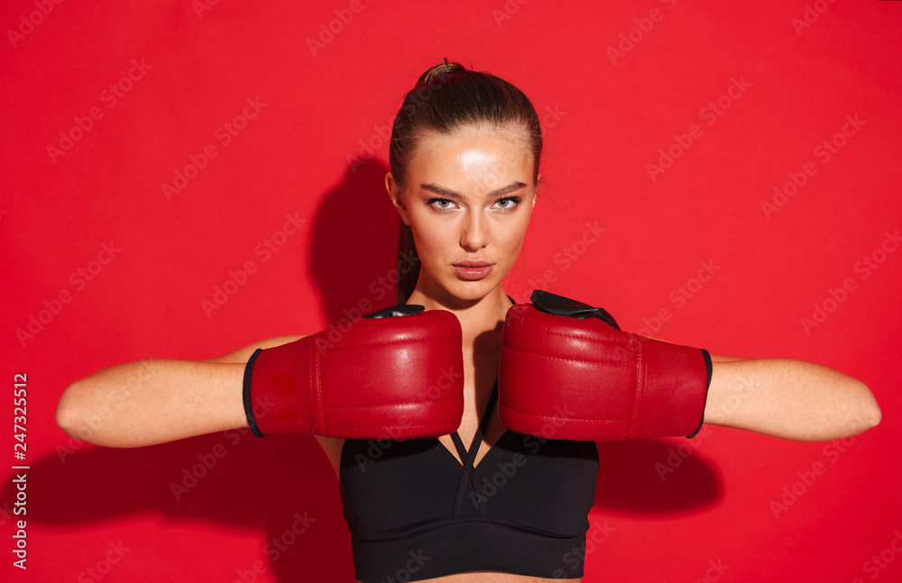 Beautiful young fitness boxer woman posing isolated over red wall background make exercises in gloves.