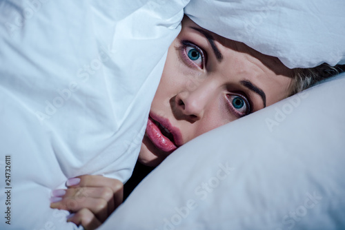 scared woman hiding behind blanket and looking at camera in bedroom at home photo