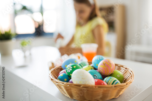 easter, holidays and people concept - colored eggs in wicker basket and girl on background at home