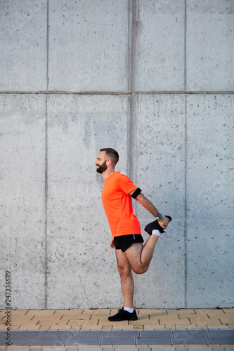 Sporty man in black and orange sportswear stretching leg and standing next to wall on the street.