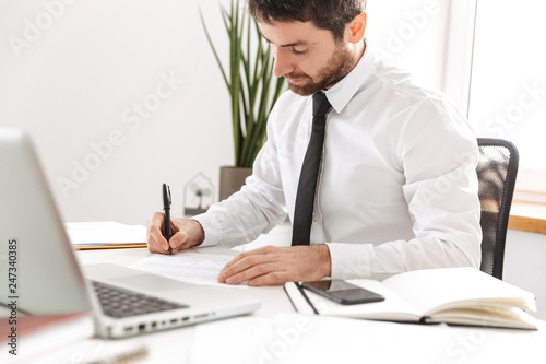 Image of concentrated office worker 30s wearing white shirt working with laptop and notebook, in modern workplace