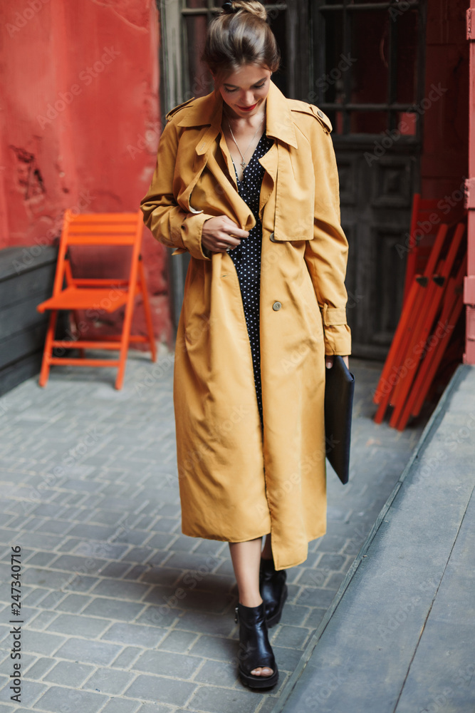 Young beautiful smiling woman in trench coat and boots holding bag in hand while spending time in old cozy courtyard Young beautiful smiling woman in trench coat and boots holding bag in hand while spending time in old cozy courtyard