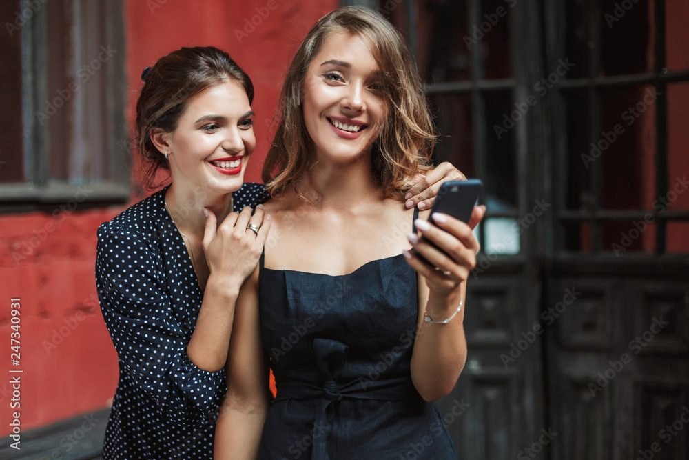 Young beautiful smiling woman in black dress holding cellphone in hand happily looking in camera with cheerful friend near in courtyard