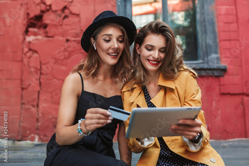 Young pretty woman in black hat with wireless earphones holding credit card in hand with cheerful woman in orange trench coat holding the tablet happily while spending time together in old courtyard