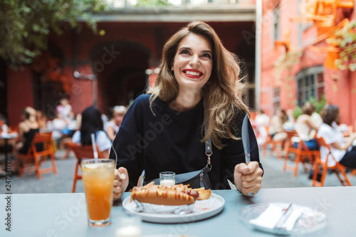 Young cheerful woman in black dress holding fork and knife in hands with food on table joyfully looking aside while spending time in cozy courtyard of cafe