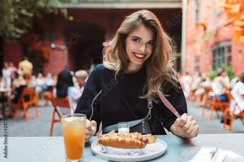 Young beautiful smiling woman in black dress holding fork and knife in hands with food on table happily looking in camera while spending time in cozy courtyard of cafe