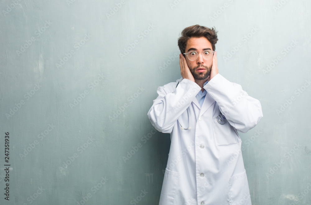 Young friendly doctor man against a grunge wall with a copy space covering ears with hands, angry and tired of hearing some sound Young friendly doctor man against a grunge wall with a copy space covering ears with hands, angry and tired of hearing some sound